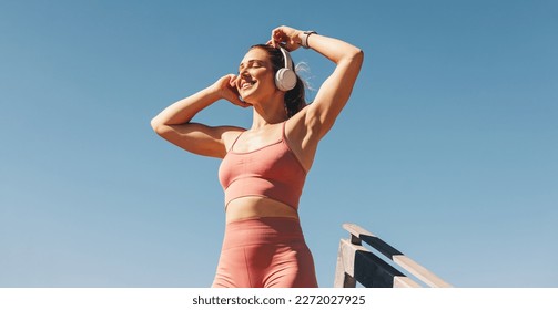 Caucasian sports woman listening to music on headphones outdoors. Woman in sportswear standing against the sky. Happy female athlete taking a break from her workout.