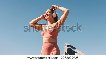 Caucasian sports woman listening to music on headphones outdoors. Woman in sportswear standing against the sky. Happy female athlete taking a break from her workout.