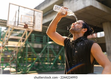 Caucasian sportsman basketball player drinking and pouring water from a bottle on his face after do sport training and playing streetball on outdoors court under highway in the city in sunny day. - Powered by Shutterstock