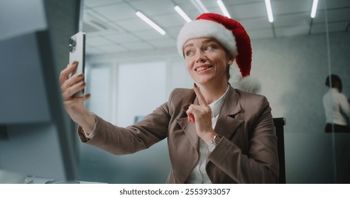 Caucasian successful businesswoman in Christmas hat sits at the workplace and talks to business partner on video call using smartphone. Female marketing specialist works in modern office. E-commerce. - Powered by Shutterstock