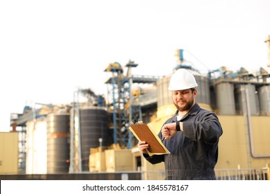 Caucasian Supervisor Looking At Watch And Holding Papers Notes Near Factory Outside. Man Wearing Helmet And Work Jumpsuit. Concept