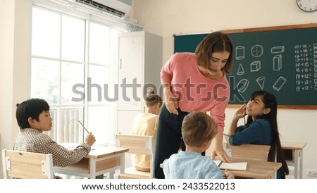 Caucasian teacher walking and check student homework at classroom while diverse children doing classwork at classroom. Diverse smart student lecturing and taking a note at elementary school. Pedagogy.