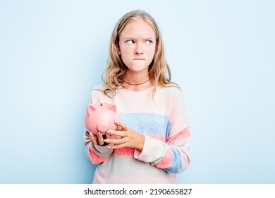 Caucasian Teen Girl Holding A Piggybank Isolated On Blue Background Confused, Feels Doubtful And Unsure.