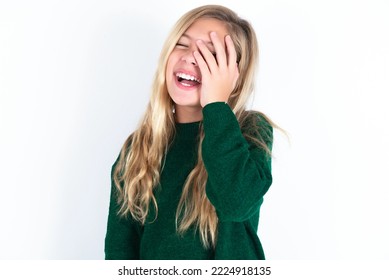 Caucasian Teen Girl Wearing Green Knitted Sweater Over White Background Makes Face Palm And Smiles Broadly, Giggles Positively Hears Funny Joke Poses