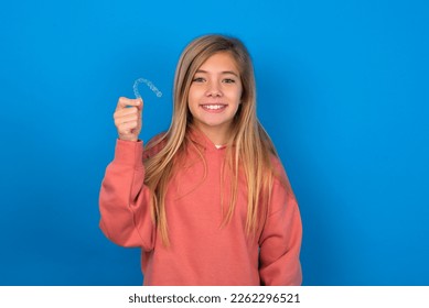 caucasian teen girl wearing pink sweater over blue wall holding an invisible braces aligner, recommending this new treatment. Dental healthcare concept. - Shutterstock ID 2262296521