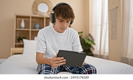 A caucasian teenage boy focused on a tablet while sitting in his bedroom, wearing headphones and pajamas. - Powered by Shutterstock