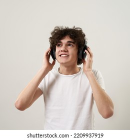 Caucasian Teenage Boy Listening Music In Headphones. Curly Guy Of Zoomer Generation Wearing T-shirt. Concept Of Modern Youngster Lifestyle. Isolated On White Background. Studio Shoot. Copy Space