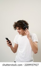 Caucasian teenage boy watching smartphone and listening music in headphones. Curly guy of zoomer generation. Concept of modern youngster lifestyle. Isolated on white background in studio. Copy space - Shutterstock ID 2209937927