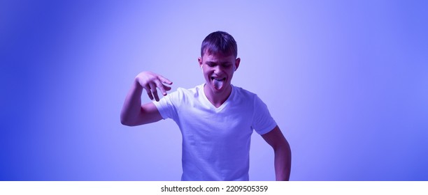 Caucasian Teenage Boy Of Zoomer Generation Sticking Out Tongue During Listening Music In Earphoness And Dancing. Modern Youngster Lifestyle. Blue With White Light Background. Studio Shoot. Copy Space