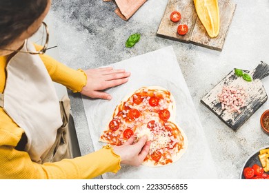 Caucasian teenage girl in an apron pouring grated cheese on pizza hearts for valentine's day with ingredients on the table, close-up side view. Concept of cooking pizza for valentine's day. - Powered by Shutterstock