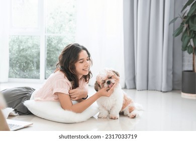 Caucasian teenage girl playing with shih tzu puppy dog at home with love. Young beautiful woman lying on floor, feeding food to little fluffy dog pet while using laptop. Small dog licking owner hand - Powered by Shutterstock