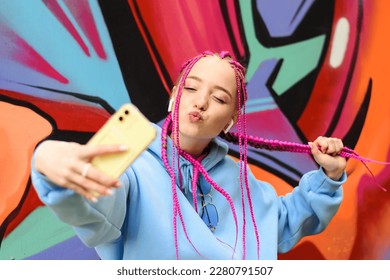 Caucasian teenage hipster girl with pink braids is using a smartphone against the background of a multicolored street wall.Summer concept.Generation Z style.Social media concept. - Powered by Shutterstock