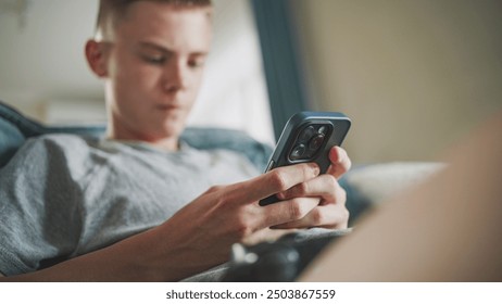 Caucasian teenager chats in messaging app with friends or watches content using mobile phone while sitting on sofa in living room. Teen boy spending weekend at home. Lifestyle concept. Close Up. - Powered by Shutterstock