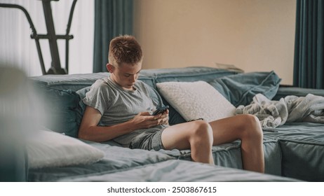 Caucasian teenager sits on sofa in cozy living room, chats with friends or watches content in social network using mobile phone. Teenage boy resting and spending weekend at home. Lifestyle concept.