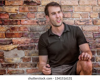 Caucasian Thirty Year Old Man Squatting And Leaning Against A Brick Wall Making A Fun Face.