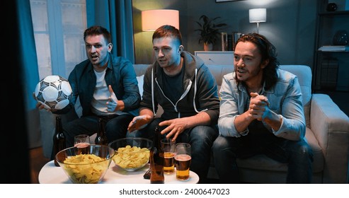 Caucasian three men best friends resting together late in evening on couch and watching sport TV while eating snacks and playing on phone. Guys sitting on sofa at home in front of television. - Powered by Shutterstock