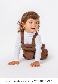 Caucasian toddler 2 years old with long curly hair on a white background in brown jumpsuit and a white turtleneck crawls on his knees on the floor on a white background. The head is turned to the side - Shutterstock ID 2232843727
