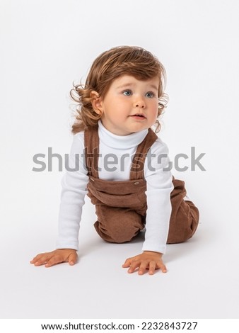 Caucasian toddler 2 years old with long curly hair on a white background in brown jumpsuit and a white turtleneck crawls on his knees on the floor on a white background. The head is turned to the side
