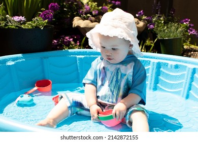 Caucasian Toddler Baby In A Hat, Playing In The Kiddie Pool On A Sunny Summer Day. Sun Protection Baby Swimwear