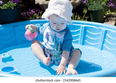 Caucasian Toddler Baby In A Hat, Playing In The Kiddie Pool On A Sunny Summer Day. Sun Protection Baby Swimwear	