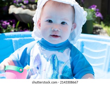 Caucasian Toddler Baby In A Hat, Playing In The Kiddie Pool On A Sunny Summer Day. Sun Protection Baby Swimwear	