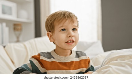 Caucasian toddler boy with blond hair sitting on a white bed in a bright bedroom, looking thoughtful - Powered by Shutterstock