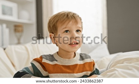 Caucasian toddler boy with blond hair sitting on a white bed in a bright bedroom, looking thoughtful