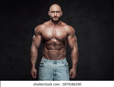 Caucasian Topless Guy With Bald Head And Muscular Build In Jeans Shorts Poses In Dark Background Looking At Camera.