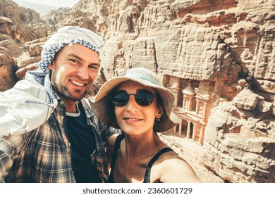 Caucasian tourist couple smile have fun enjoy Petra ancient city over Treasury or Al-khazneh take smartphone photo together on holiday vacation. UNESCO World Heritage site.