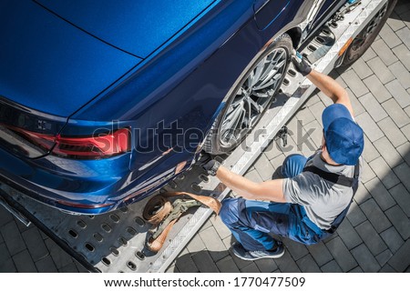 Caucasian Towing Truck Driver in His 40s Preparing Vehicle For Car Dealership Customer. Brand New Car Home Delivery on Towing Truck.