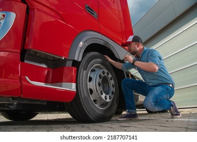 Caucasian Trucker Closely Checking the Condition of the Tires in His Large Heavy Duty Red Semi Truck Before Setting Off to Deliver a Cargo. Transportation and Logistics Workers Theme. - Shutterstock ID 2187707141