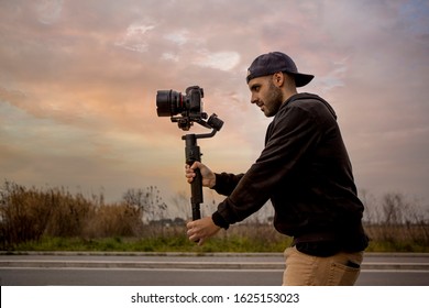 Caucasian videographer filming with cinema gimbal video dslr at sunset , professional video, video maker in event. Cinema lens on gimbal. Medium shot from left - Shutterstock ID 1625153023