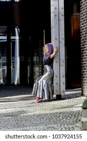 Caucasian White Female Model Standing On The Lobby Of The Building And Enjoys Weather. Beautiful Woman With Long Purple Hair, In A Gray Dress Resting In The City, Modern Fashion Concept.
