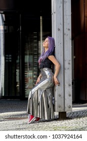 Caucasian White Female Model Standing On The Lobby Of The Building And Enjoys Weather. Beautiful Woman With Long Purple Hair, In A Gray Dress Resting In The City, Modern Fashion Concept.
