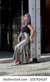 Caucasian White Female Model Standing On The Lobby Of The Building And Enjoys Weather. Beautiful Woman With Long Purple Hair, In A Gray Dress Resting In The City, Modern Fashion Concept.
