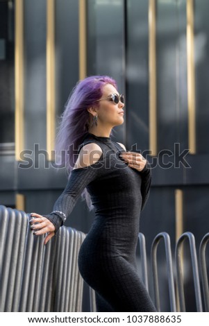 Caucasian white female model standing on the balcony of the building and enjoys weather. Beautiful woman with long purple hair, in a gray dress resting in the city, modern fashion concept.