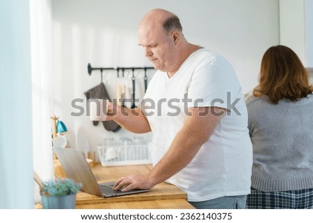 Caucasian - white oversize obese man checking an emails and messages on laptop - notebook computer on the counter top in kitchen between drinks a cup of coffee in morning.