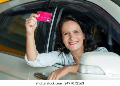 Caucasian woman adult leaning out of car window smiling holding up credit card. light denim jacket. Indicates satisfaction after completing a car service or insurance transaction at a repair shop. - Powered by Shutterstock