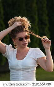 Caucasian woman with afro curls hairstyle posing standing at sunrise in summertime heat. Blond girl in sunwear and white sundress fixing hairdo. Warm sunny day. Sunshine