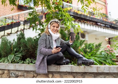 Caucasian Woman In Autumn Coat Listens To Music On Headphones And Looks At Mobile Phone Outside