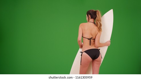 Caucasian woman in bikini shown from behind holding surfboard on green screen