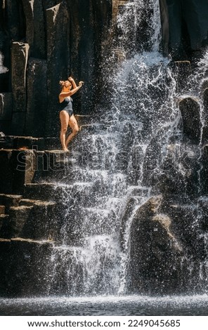 Caucasian woman in black swimsuit refreshing under the falling water streams flowing on black volcanic stone cascades. Rochester Falls waterfall - popular tourist spot in Savanne district in Mauritius