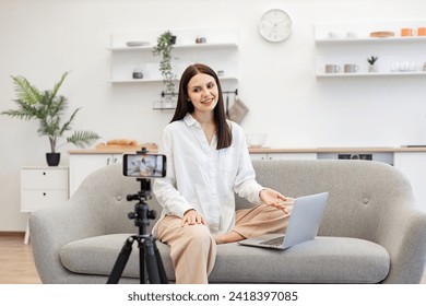 Caucasian woman with brunette hair reading information from modern laptop during live broadcast on social network. Demanding tutor using modern smartphone with tripod for online training. - Powered by Shutterstock