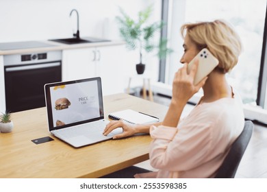 Caucasian woman in casual attire using laptop to order food online. She holds phone to her ear while browsing food delivery website. Bright morning light fills modern kitchen with minimalistic decor. - Powered by Shutterstock