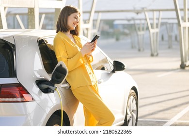 Caucasian Woman Charging An Electric Car At A Charging Station Near An Electric Solar Power Plant, Using Her Smartphone While Waiting.