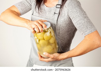 A caucasian woman chef wearing apron is trying to open a stubborn jar lid. She uses force to unscrew the lid in kitchen. Concept image for pickling tomatoes and gherkins and difficulty in opening lids