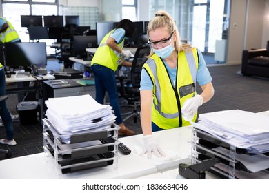 Caucasian Woman And Colleagues Wearing Hi Vis Vests, Gloves, Safety Glasses And Face Masks Sanitizing An Office Using Disinfectant. Hygiene In Workplace During Coronavirus Covid 19 Pandemic.