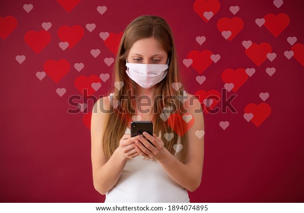 Caucasian woman dating online through internet\
during coronavirus pandemic with hearts around on red background.\
Valentine with covid 19 disease. Girlfriend waring face mask and\
typing on her phone.