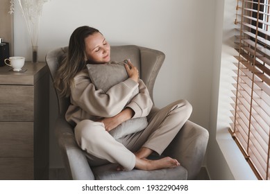 Caucasian woman dressed in comfy grey loungewear sitting in a cozy armchair in her bedroom and holding a decorative pillow. Coziness and comfort concept