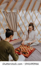 Caucasian Woman Dressed In White Dress And Man Sitting In A Yurt And Playing Chess Enjoying Table Games. White Yurt Tent In Background. Adventure Luxury Accommodation. Tent Yurt Hotel Stay For Family